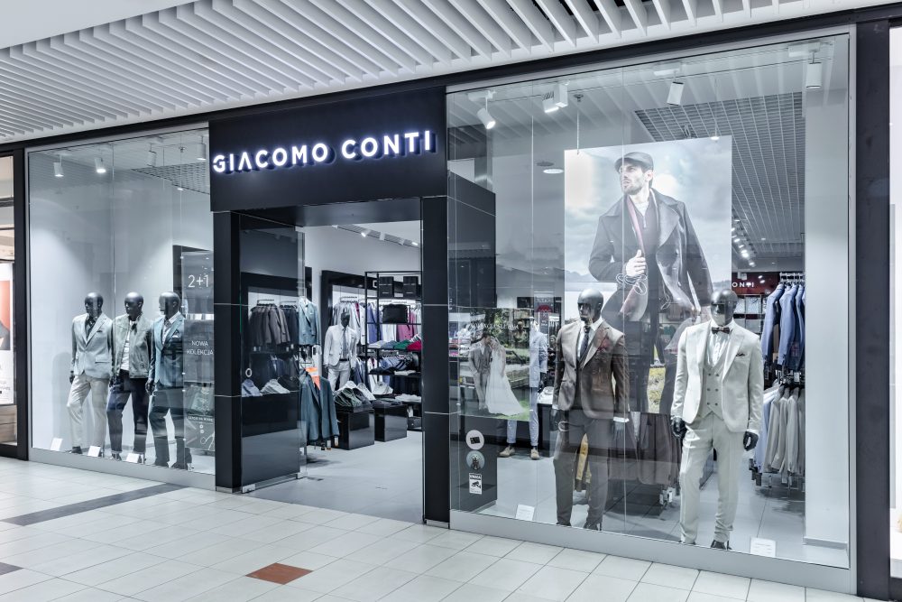 Elegancki front salonu Giacomo Conti w Pasażu Grunwaldzkim. Nad wejściem widoczne stylowe logo marki. Witryny prezentują manekiny w nienagannie skrojonych garniturach, a ciepłe oświetlenie wnętrza zachęca do wejścia.