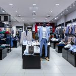asne i luksusowe wnętrze salonu Giacomo Conti. Na drewnianych i metalowych regałach starannie poukładane są koszule, marynarki oraz akcesoria, takie jak krawaty i poszetki. W głębi widać przestronne lustra i strefę dopasowania odzieży.
