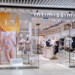 Frontowy widok na wejście do salonu Intimissimi. Nad wejściem widnieje klasyczne, minimalistyczne logo marki. Witryny są zaaranżowane w subtelny sposób, prezentując najnowszą kolekcję bielizny i odzieży domowej na eleganckich manekinach.