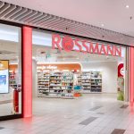 Frontowy widok na wejście do drogerii Rossmann. Nad wejściem znajduje się duże, czerwone logo.
