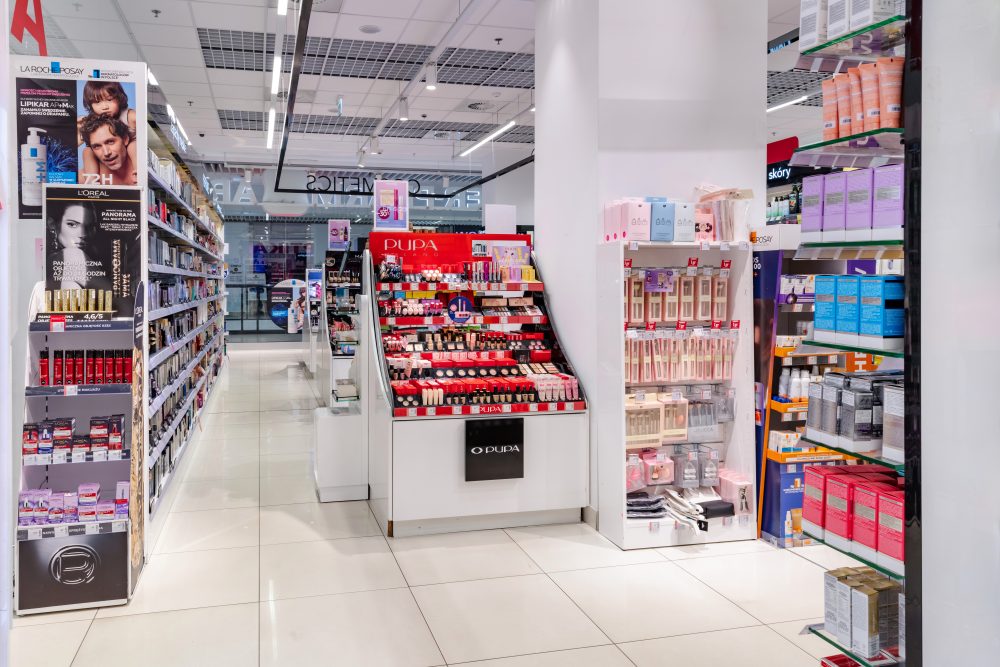 Wnętrze działu kosmetycznego w Super-Pharm. Widoczne są rzędy szaf z markami makijażowymi oraz regały z produktami do pielęgnacji twarzy.