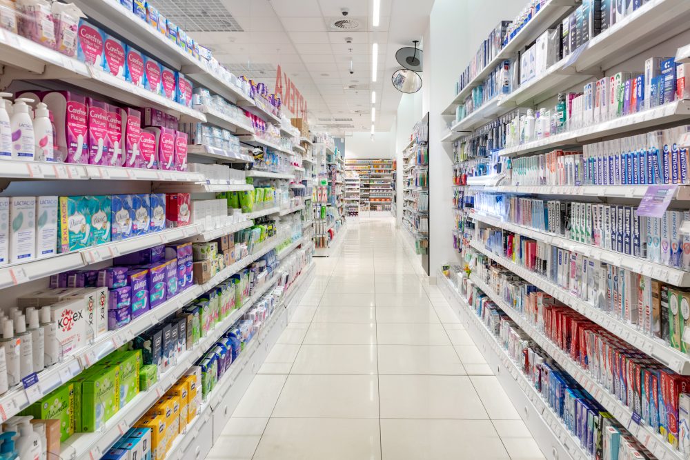 Widok na szeroką i uporządkowaną alejkę w Super-Pharm. Na regałach po jednej stronie widoczne są pasty do zębów i artykuły do higieny jamy ustnej, a po drugiej produkty higieny intymnej i podpaski.