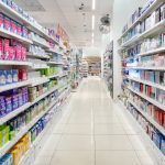 Widok na szeroką i uporządkowaną alejkę w Super-Pharm. Na regałach po jednej stronie widoczne są pasty do zębów i artykuły do higieny jamy ustnej, a po drugiej produkty higieny intymnej i podpaski.