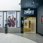 Wejście do klubu fitness Zdrofit w galerii handlowej. Po prawej stronie widoczne jest otwarte, podświetlone wejście z podświetlanym logo ZDROFIT na czarnym tle.
