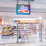 Zbliżenie na nowoczesne wejście do drogerii dm w Pasażu Grunwaldzkim. Nad wejściem widoczne kolorowe logo marki. W witrynie wyeksponowane są najnowsze gazetki promocyjne oraz produkty marek własnych, takich jak Balea czy Alverde.