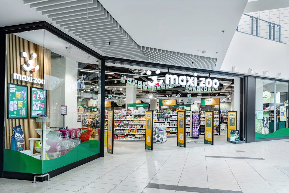 Perspektywa boczna na wejście i witrynę sklepu Maxi Zoo w pasażu handlowym. Widoczne są plakaty informacyjne o promocjach oraz przejrzysty układ wejścia, który ułatwia komunikację wewnątrz galerii.