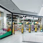 Perspektywa boczna na wejście i witrynę sklepu Maxi Zoo w pasażu handlowym. Widoczne są plakaty informacyjne o promocjach oraz przejrzysty układ wejścia, który ułatwia komunikację wewnątrz galerii.