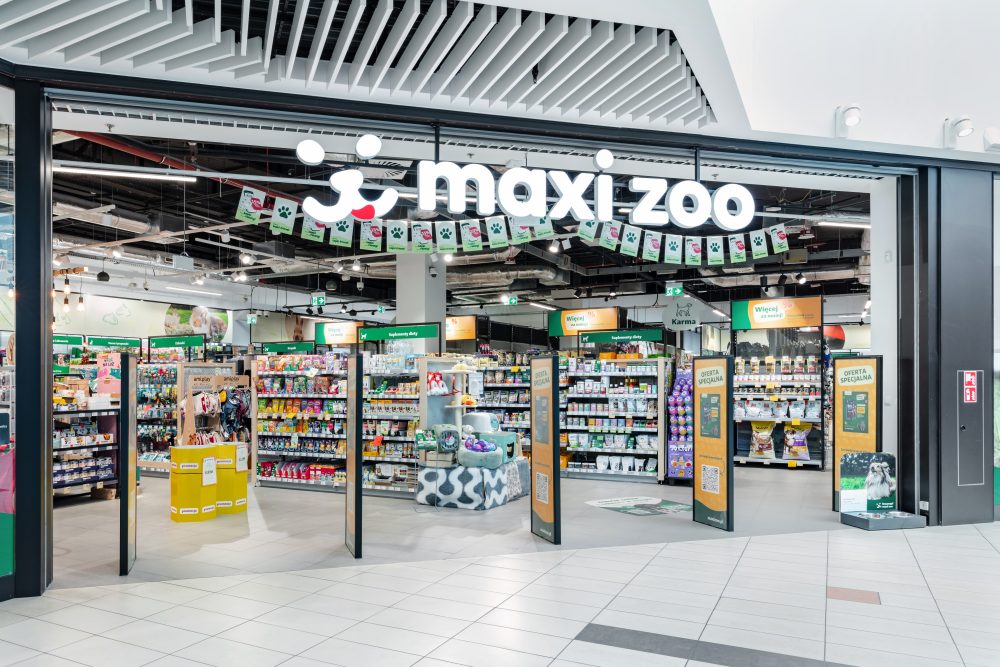 Frontowy widok na duże, otwarte wejście do sklepu Maxi Zoo. Nad wejściem znajduje się charakterystyczne logo z sylwetkami psa i kota na czerwonym tle.