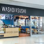 Widok na elegancki front restauracji Wasabi Sushi w strefie gastronomicznej. Nad wejściem widnieje charakterystyczne logo marki.