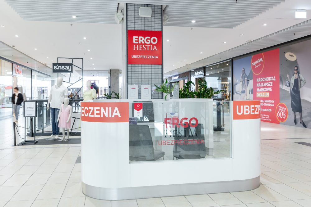 Nowoczesna wyspa handlowa Ergo Hestia usytuowana w pasażu galerii handlowej. Punkt jest biało-niebieski, z wyraźnym logo marki oraz napisami informującymi o rodzajach ubezpieczeń (auto, dom, podróż). Przy ladzie widoczne są stanowiska obsługi z monitorami i wygodne miejsca dla klientów.