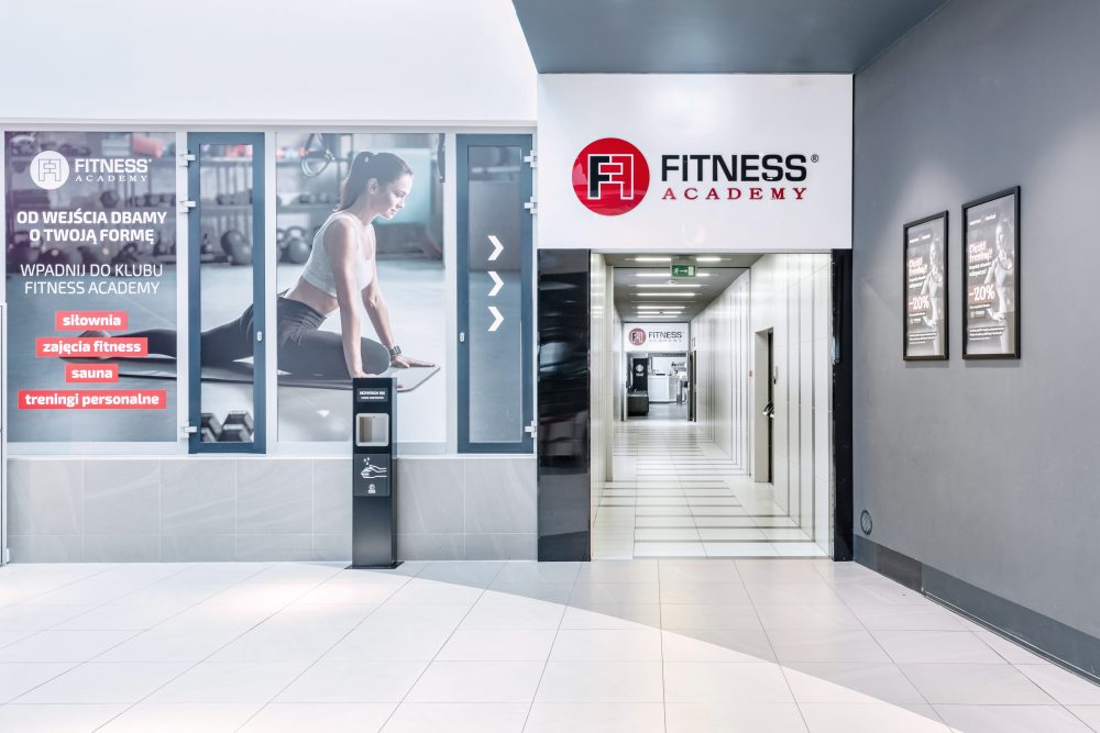 Nowoczesne wejście do klubu Fitness Academy. Widoczne duże, podświetlane logo marki oraz przeszklone drzwi prowadzące do recepcji. Design jest dynamiczny, utrzymany w sportowej stylistyce.