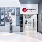 Nowoczesne wejście do klubu Fitness Academy. Widoczne duże, podświetlane logo marki oraz przeszklone drzwi prowadzące do recepcji. Design jest dynamiczny, utrzymany w sportowej stylistyce.