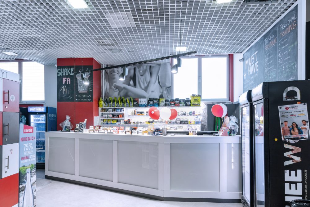Estetyczna lada recepcyjna w klubie fitness. Za ladą widoczny logotyp Fitness Academy. Obok znajdują się bramki wejściowe na czytnik kart oraz ekspozycja napojów izotonicznych.