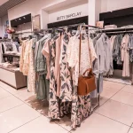 Jasne wnętrze sklepu odzieżowego BETTY BARCLAY z widoczną w centrum ekspozycją ubrań na metalowym stojaku. Na wieszakach wiszą ubrania w pastelowych, stonowanych odcieniach: wzorzysta sukienka w liście, ażurowa bluzka oraz odzież w odcieniach beżu, różu i zieleni.