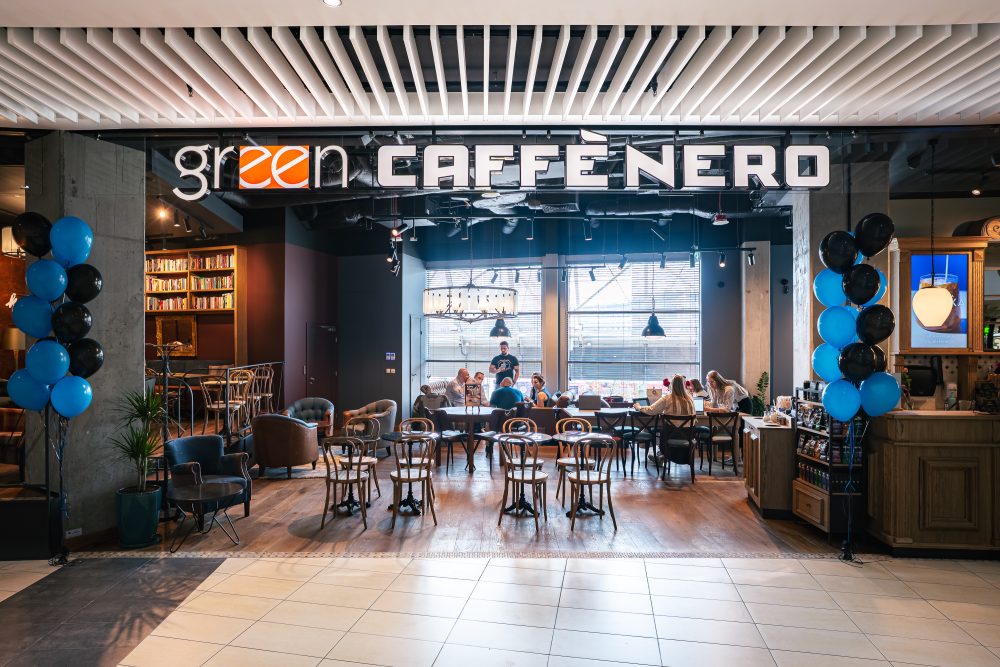 Kadr przedstawia widok na wejście do przestronnej kawiarni, nad którym umieszczono duży, podświetlany szyld z nazwą „green CAFFÈ NERO”. Po bokach przejścia znajdują się kolumny udekorowane pękami niebieskich i czarnych balonów, a w głębi lokalu widoczne są stoliki, przy których siedzą klienci.