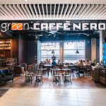 Kadr przedstawia widok na wejście do przestronnej kawiarni, nad którym umieszczono duży, podświetlany szyld z nazwą „green CAFFÈ NERO”. Po bokach przejścia znajdują się kolumny udekorowane pękami niebieskich i czarnych balonów, a w głębi lokalu widoczne są stoliki, przy których siedzą klienci.