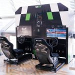 Stanowisko z dwoma profesjonalnymi symulatorami wyścigowymi SIMNATION ustawione w przestrzeni galerii handlowej. Na pierwszym planie widoczne są dwa ergonomiczne fotele kubełkowe z napisem „SLIDE” oraz zamontowane przed nimi kierownice i pedały. Nad stanowiskiem znajduje się duża czarno-biało-zielona obudowa z ekranami wyświetlającymi symulację toru wyścigowego.
