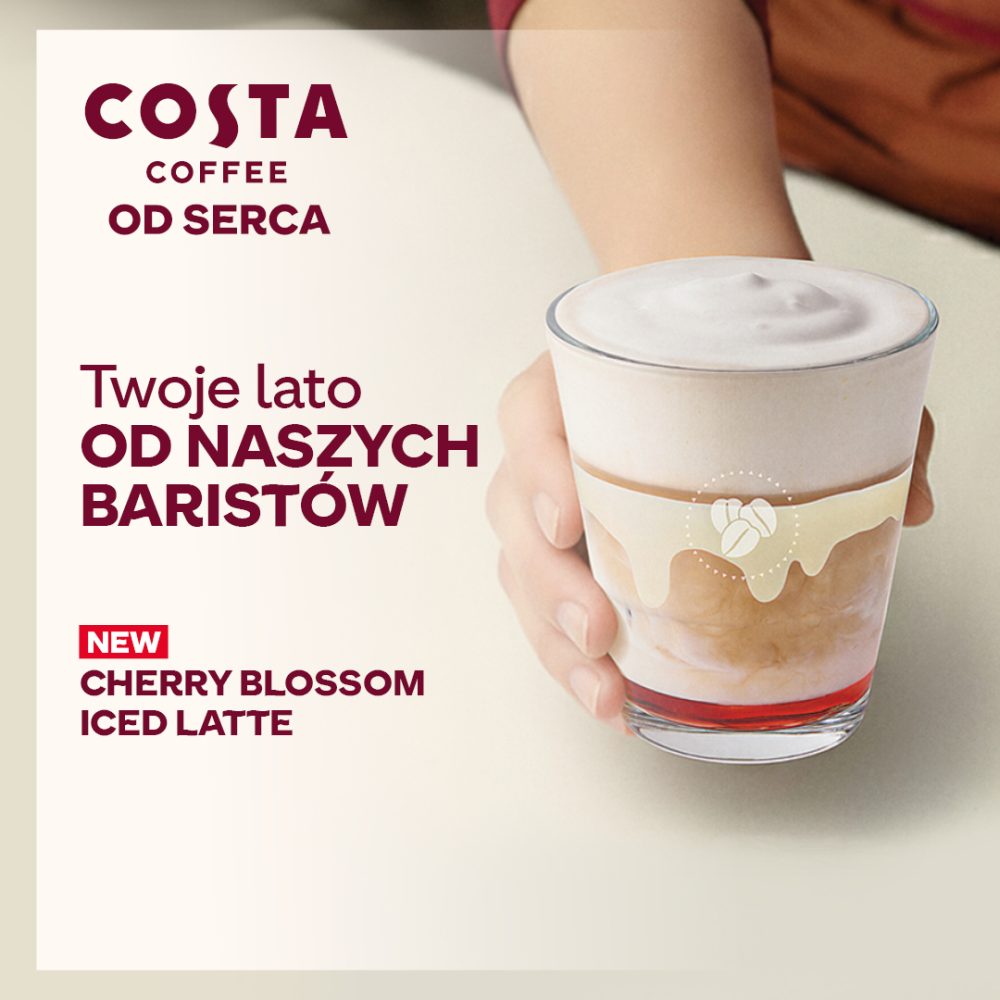 Grafika reklamowa Costa Coffee prezentuje nowość w menu – Cherry Blossom Iced Latte, czyli warstwową mrożoną kawę z czerwonym syropem na dnie. Dłoń baristy trzyma przezroczystą szklankę wypełnioną napojem z gęstą, jasną pianką, ozdobioną grafiką ziaren kawy.