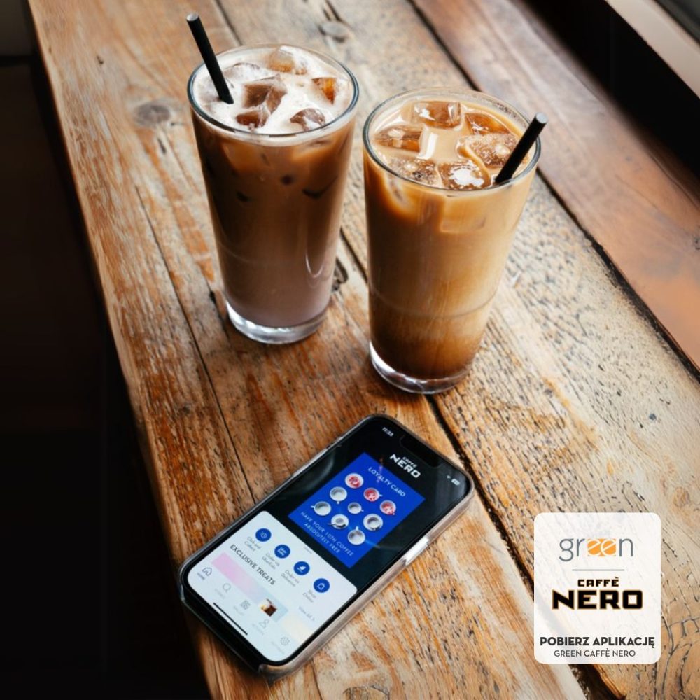 Grafika reklamowa kawiarni Green Caffè Nero przedstawia dwie wysokie szklanki mrożonej kawy z kostkami lodu i słomkami, ustawione na drewnianym stole obok smartfona z włączoną aplikacją lojalnościową. W prawym dolnym rogu umieszczono logo marki z dopiskiem „POBIERZ APLIKACJĘ Green Caffè Nero”.