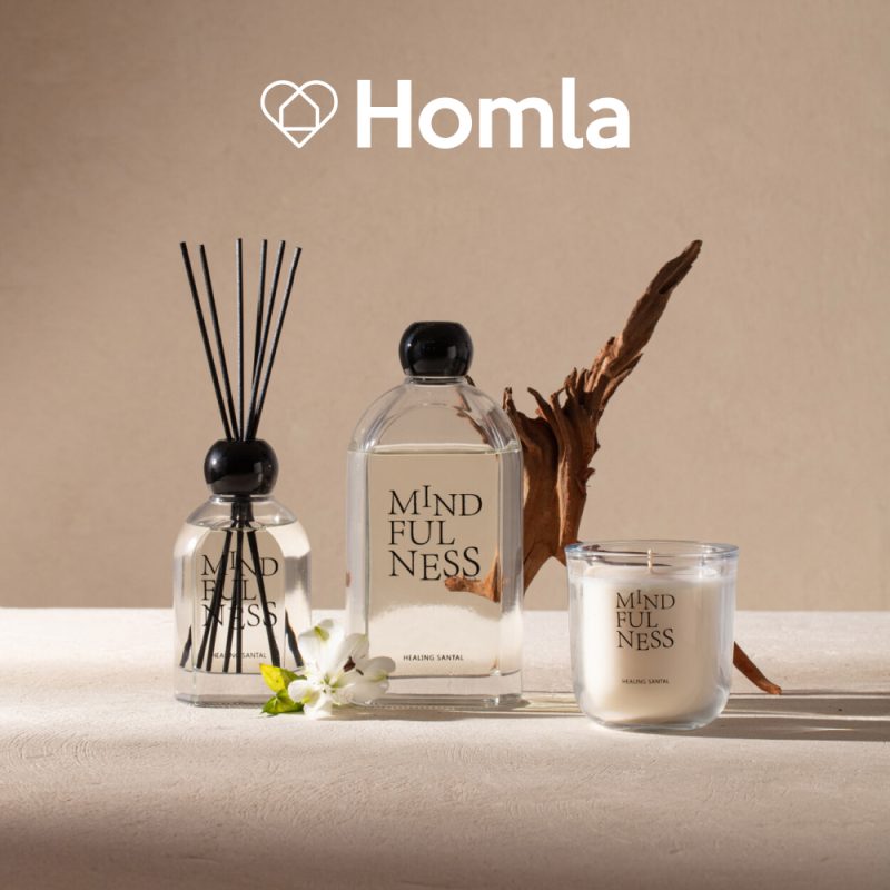 Grafika reklamowa marki Homla przedstawia zestaw produktów zapachowych z serii „Mindfulness” o zapachu „Healing Santal”, w skład którego wchodzi dyfuzor z patyczkami, butelka z płynem oraz świeca w szkle. Całość została zaaranżowana na jasnym, beżowym tle w otoczeniu naturalnych dekoracji, takich jak kawałek drewna oraz biały kwiat.