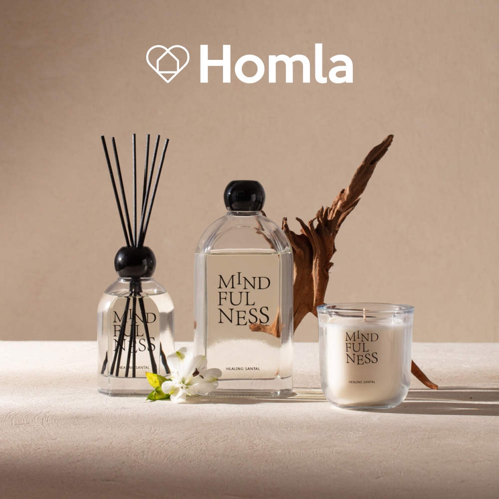 Grafika reklamowa marki Homla przedstawia zestaw produktów zapachowych z serii „Mindfulness” o zapachu „Healing Santal”, w skład którego wchodzi dyfuzor z patyczkami, butelka z płynem oraz świeca w szkle. Całość została zaaranżowana na jasnym, beżowym tle w otoczeniu naturalnych dekoracji, takich jak kawałek drewna oraz biały kwiat.