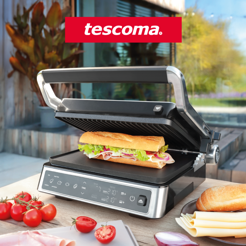 Na zdjęciu widać elektryczny grill kontaktowy marki Tescoma, na którym przygotowywana jest kanapka z bagietki, szynki, sera, sałaty i warzyw. Grill stoi na stole na zewnątrz, w tle widoczny jest nowoczesny taras i ogród. Obok urządzenia leżą świeże pomidory, ser, cebula i bagietka, co tworzy apetyczną, letnią scenę kulinarną.