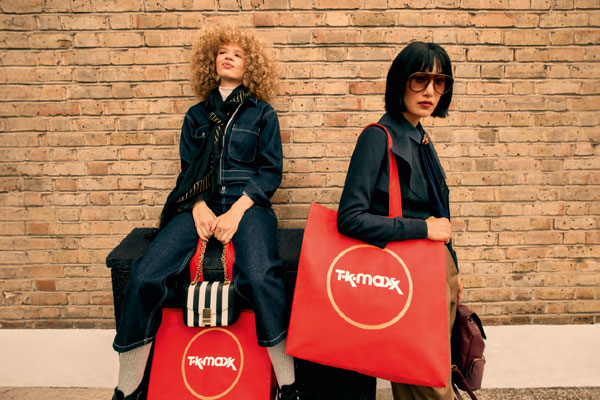 Dwie osoby prezentujące stylizacje modowe stoją i siedzą przy ceglanej ścianie, trzymając czerwone torby zakupowe TK Maxx.