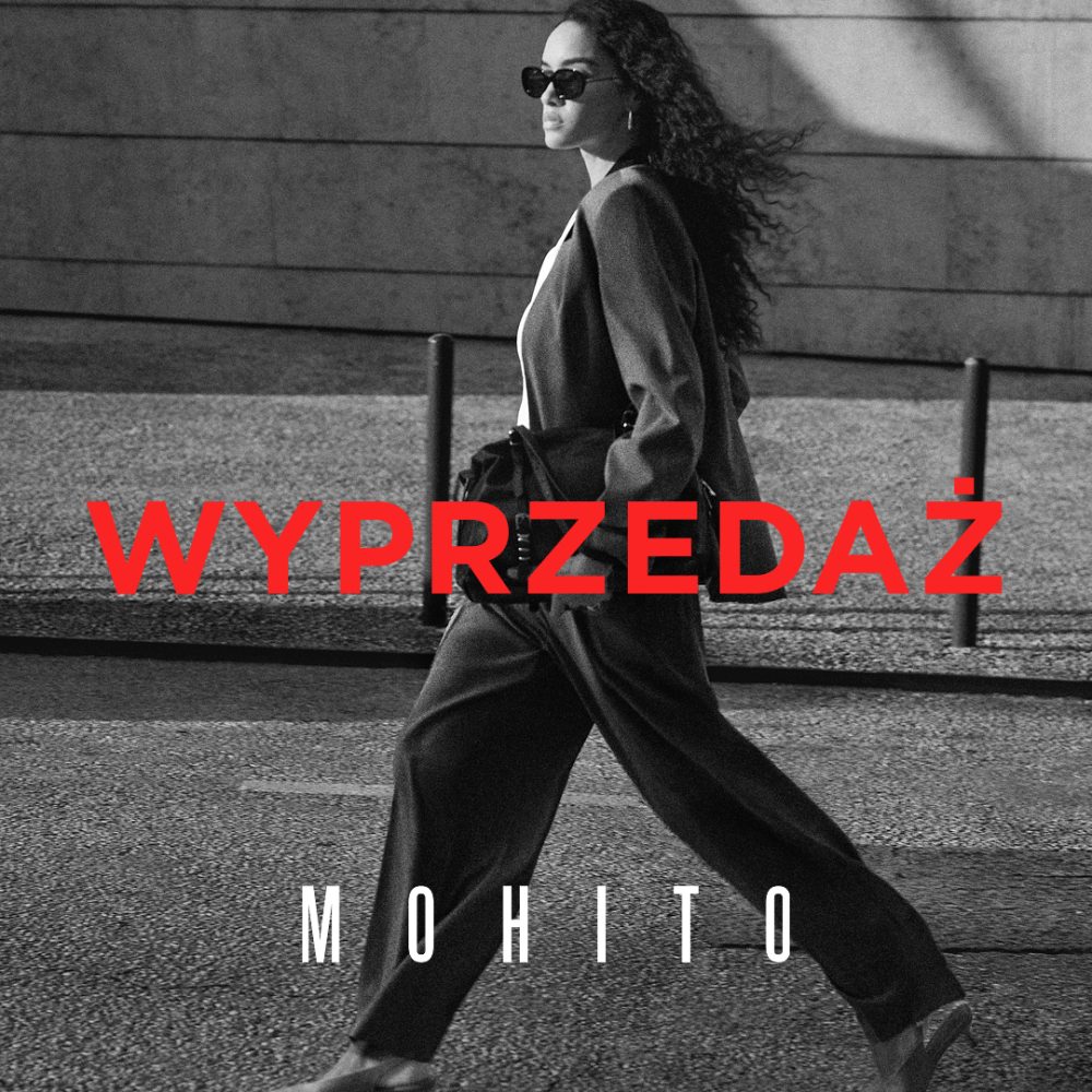 Czarno-białe zdjęcie wizerunkowe marki Mohito przedstawia idącą ulicą kobietę w eleganckim garniturze i okularach przeciwsłonecznych, trzymającą torebkę pod pachą. Na pierwszym planie wyróżnia się duży, jaskrawoczerwony napis „WYPRZEDAŻ”, a pod nim umieszczono białe logo marki „MOHITO”.