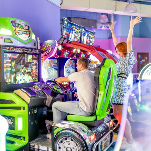 Mężczyzna grający w wyścigi samochodowe na automacie w salonie gier, któremu kibicuje uśmiechnięta kobieta z uniesionymi rękami. W tle kolorowe automaty arcade i neonowe oświetlenie.