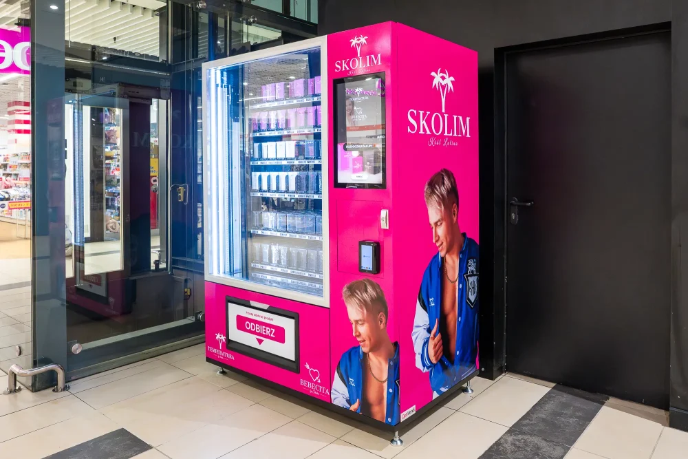 Różowy automat vendingowy sygnowany marką Skolim w galerii handlowej, wypełniony kosmetykami i perfumami. Na obudowie widnieją wizerunki wokalisty oraz logo z palmami.