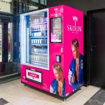 Różowy automat vendingowy sygnowany marką Skolim w galerii handlowej, wypełniony kosmetykami i perfumami. Na obudowie widnieją wizerunki wokalisty oraz logo z palmami.