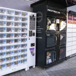 Trzy automaty vendingowe stojące na zewnątrz budynku. Po lewej stronie znajduje się biały automat z przezroczystymi, numerowanymi skrytkami zawierającymi drobne produkty i puszki.