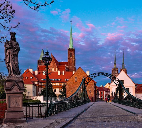 Widok na zabytkowy, stalowy Most Tumski we Wrocławiu o zmierzchu. Po lewej stronie pierwszego planu stoi kamienna rzeźba na postumencie, a na wprost rozciąga się brukowa droga prowadząca w stronę Ostrowa Tumskiego. W tle widoczne są ceglane budynki sakralne i wieże kościołów na tle malowniczego nieba w odcieniach fioletu, różu i błękitu.