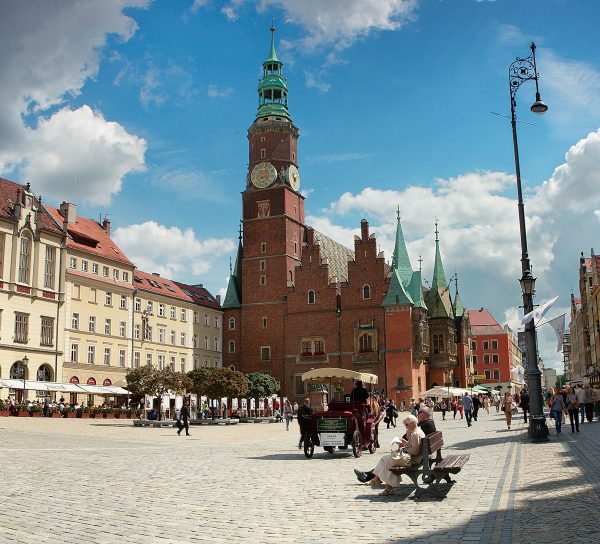Słoneczny, brukowany rynek starego miasta z gotyckim ratuszem o wysokiej wieży z zegarem i zielonym hełmem. Na pierwszym planie po prawej stronie na ławce siedzą ludzie, obok przejeżdża konny dorożka, a po placu spacerują przechodnie. Po lewej stronie widoczne są kolorowe kamienice z ogródkami restauracyjnymi, a nad całością rozciąga się niebieskie niebo z białymi chmurami.