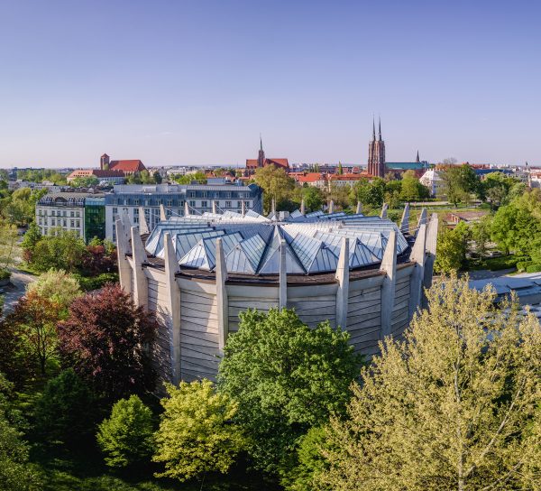 Widok z góry na rotundę Panoramy Racławickiej we Wrocławiu, otoczoną gęstą zielenią parkowych drzew. Betonowy budynek o charakterystycznej, gwiaździstej konstrukcji dachu usytuowany jest w centrum kadru, a w tle rozpościera się panorama miasta z widocznymi wieżami kościołów na Ostrowie Tumskim pod pogodnym niebem.