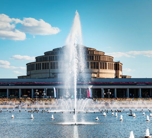 Wysoki strumień wody z Fontanny Multimedialnej strzelający w górę na pierwszym planie wrocławskiego kompleksu Hali Stulecia. W tle znajduje się monumentalna, modernistyczna Hala Stulecia ze swoją charakterystyczną kopułą, a przed nią rozciąga się kolumnada pergoli i basen z mniejszymi wodotryskami pod jasnym, słonecznym niebem.